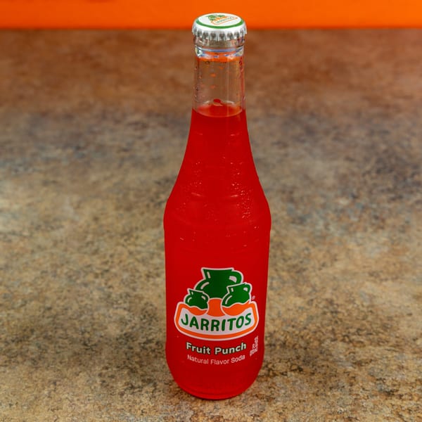 A cold bottle of red Jarritos Fruit Punch soda with condensation, on a textured brown surface.