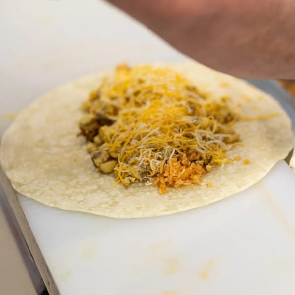A flour tortilla filled with seasoned meat, rice, and shredded cheese on a white cutting board.