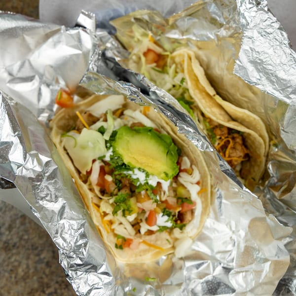 Two Burrito Bandito tacos, one topped with avocado, sour cream, and salsa, wrapped in foil.