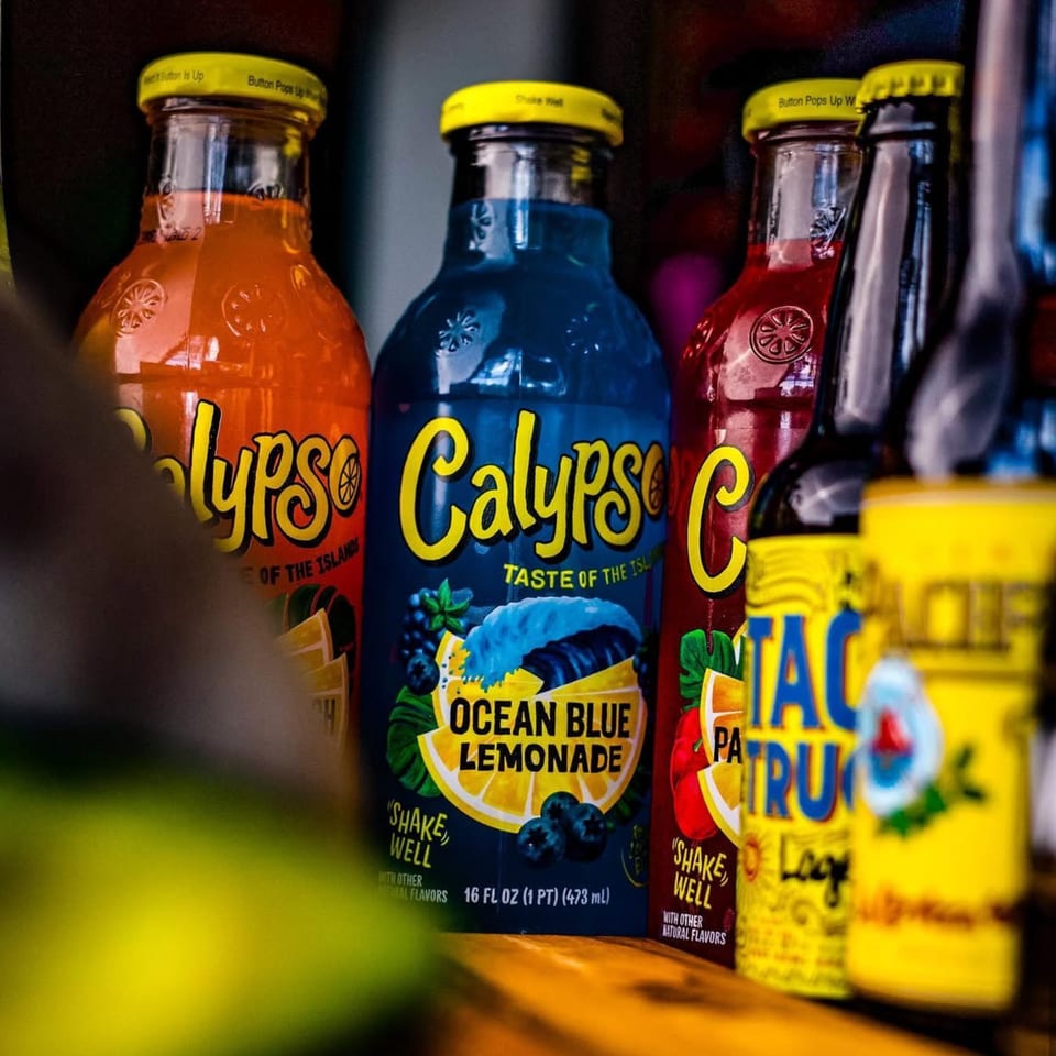 A close-up of several colorful Calypso brand lemonade bottles, including Ocean Blue, on a wooden surface.